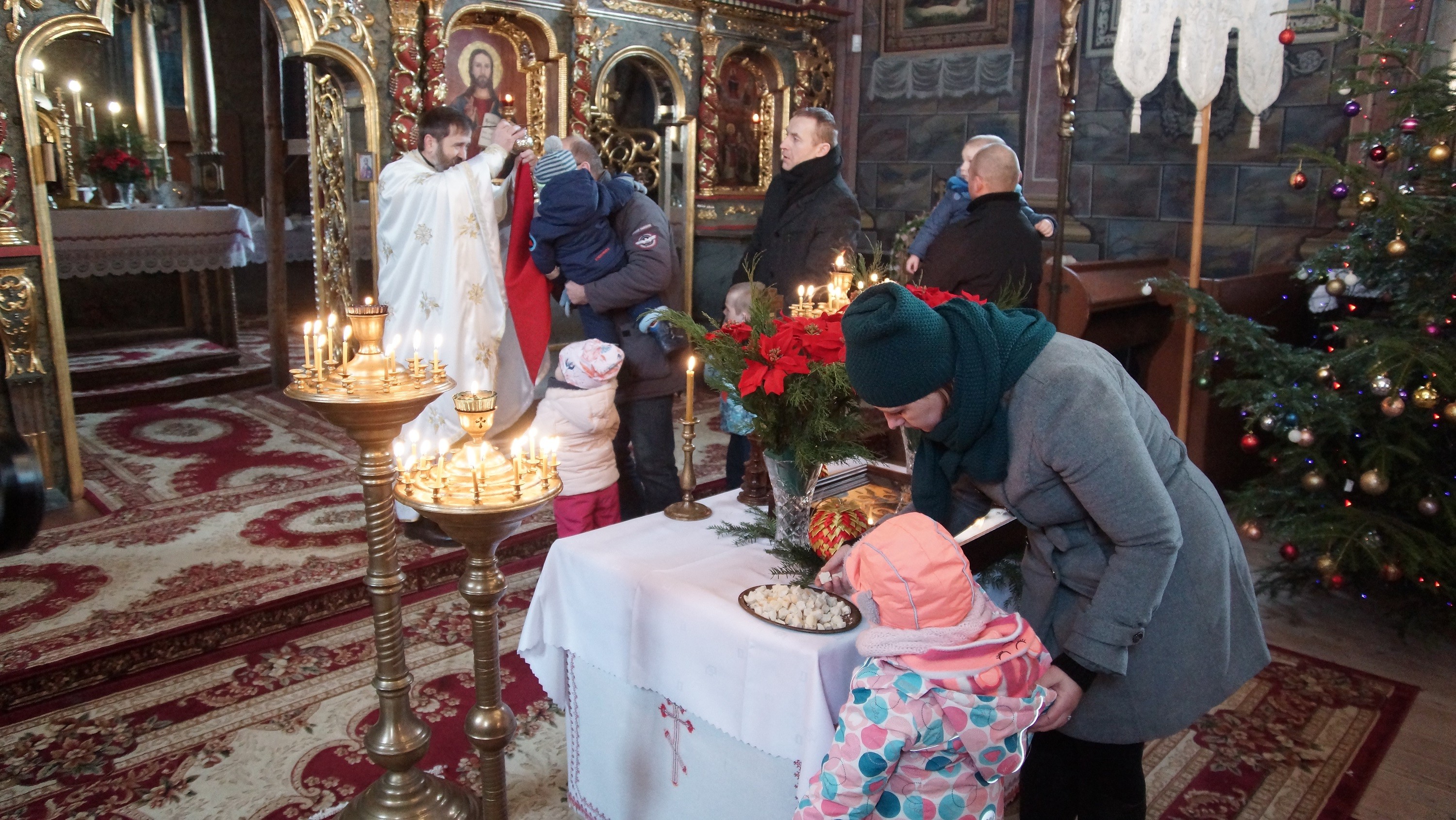 Wnętrze cerkwi. Po środku stoi niewielki stolik, a na nim znajduje się ikona Bożego Narodzenia oraz płaskie naczynie z drobnymi kawałkami chleba. Przy stoliku stoi kobieta z dzieckiem. Kobieta bierze kawałek chleba. W tle widoczny ksiądz trzymający kielich. Przed nim kolejka wiernych z dziećmi.