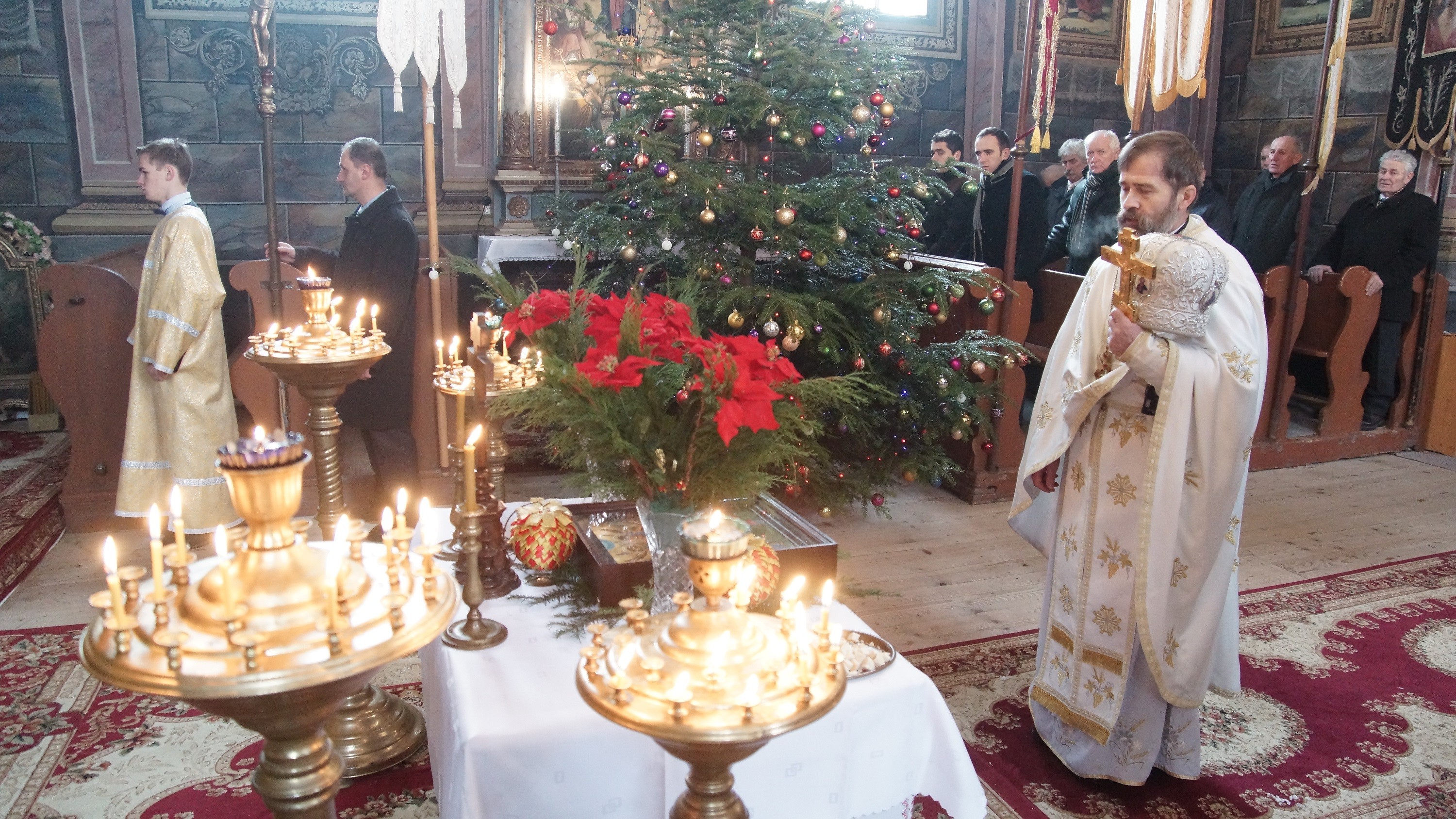 Wnętrze cerkwi. Przed niewielkim stolikiem z ikoną Bożego Narodzenia stoi ksiądz. W dłoni trzyma krzyż oraz mitrę - nakrycie głowy. W tle ławy z wiernymi i bożonarodzeniowa choinka.