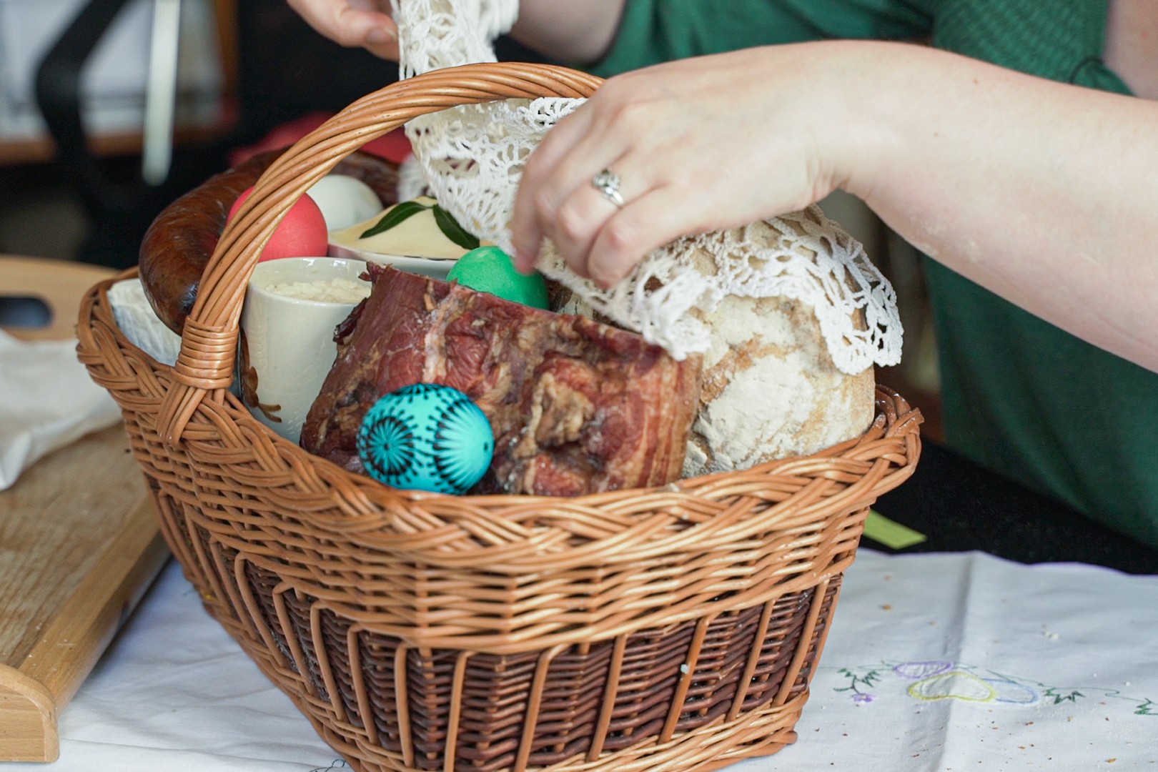 koszyczek wielkanocny, a w nim jajko, boczek, kiełbasa, chleb. Potrawy koronkową chustą okrywa kobieta